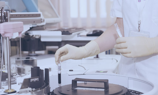 Dr. Luke Fullenkamp : A laboratory technician in a white coat and gloves placing a vial into an analytical machine in a clinical lab.