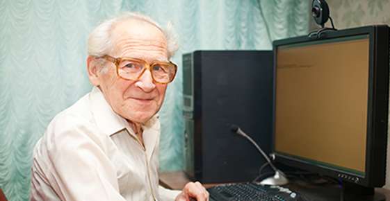 Dr. Luke Fullenkamp : Elderly man with glasses, managing macular degeneration, smiling at the camera while sitting at a desk with a computer monitor and keyboard.